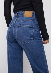 Jeans de denim azul de cintura alta con un parche de cuero marrón en la parte trasera y dos bolsillos traseros, con una textura ligeramente desteñida.
