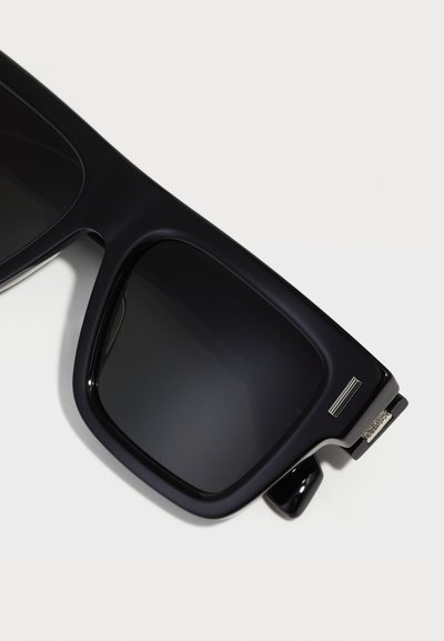 Black sunglasses with large square lenses, glossy plastic frame, silver hardware accents, and dark tinted lenses. Features unique temple design.