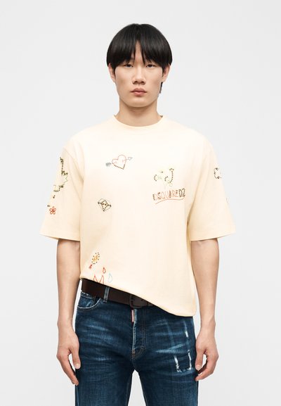 Νέος άντρας με εκρού υπερμεγέθη μπλουζάκι με κεντημένα σχέδια και μπλε τζιν με φθορές, στέκεται μπροστά σε απλό λευκό φόντο.