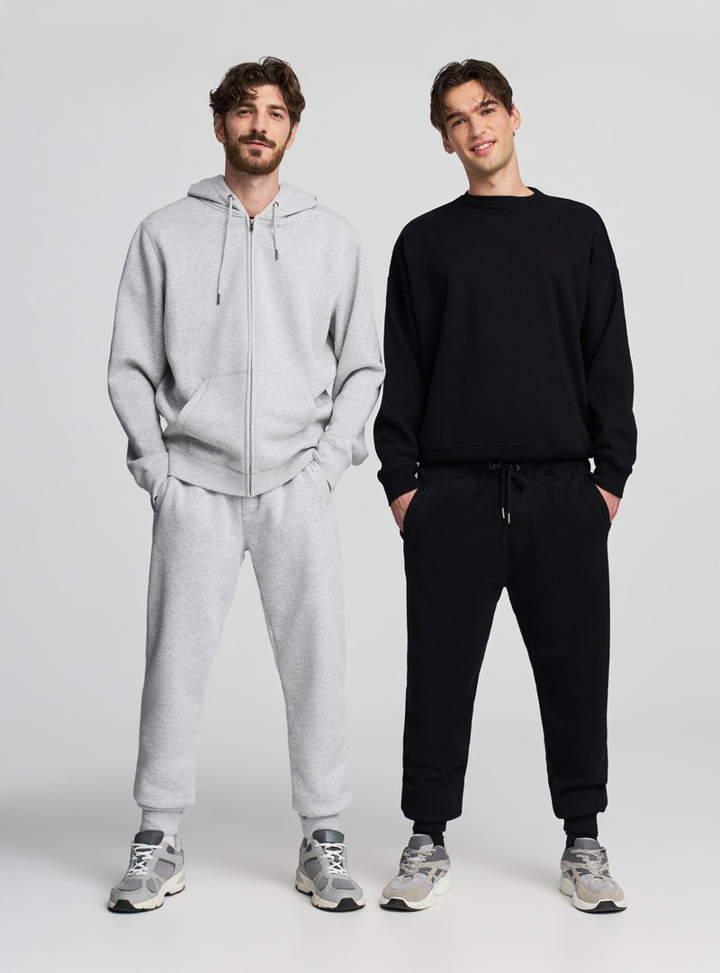 Dwóch mężczyzn nosi dopasowane casualowe stroje: jasnoszara bluza z kapturem na zamek z pasującymi spodenkami dresowymi oraz czarna bluza z okrągłym dekoltem z czarnymi joggerami.