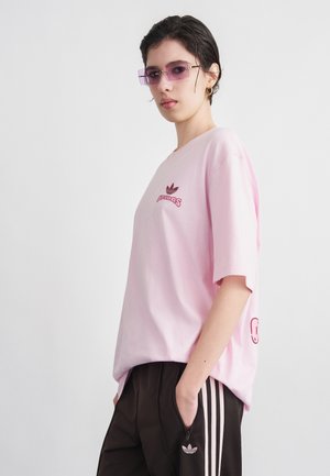 Persona joven con camiseta rosa de Adidas, pantalones deportivos negros de Adidas, gafas con cristales tintados en púrpura, de pie con las manos en los bolsillos frente a un fondo blanco.