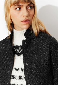 Veste noire texturée avec accents argentés sur un pull blanc avec motifs géométriques noirs. Boutons visibles, col rond.