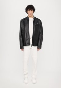 Veste en cuir noire avec col montant, fermeture éclair à l'avant et poches latérales. Portée par-dessus un t-shirt blanc, associée à un pantalon blanc et des chaussures blanches.