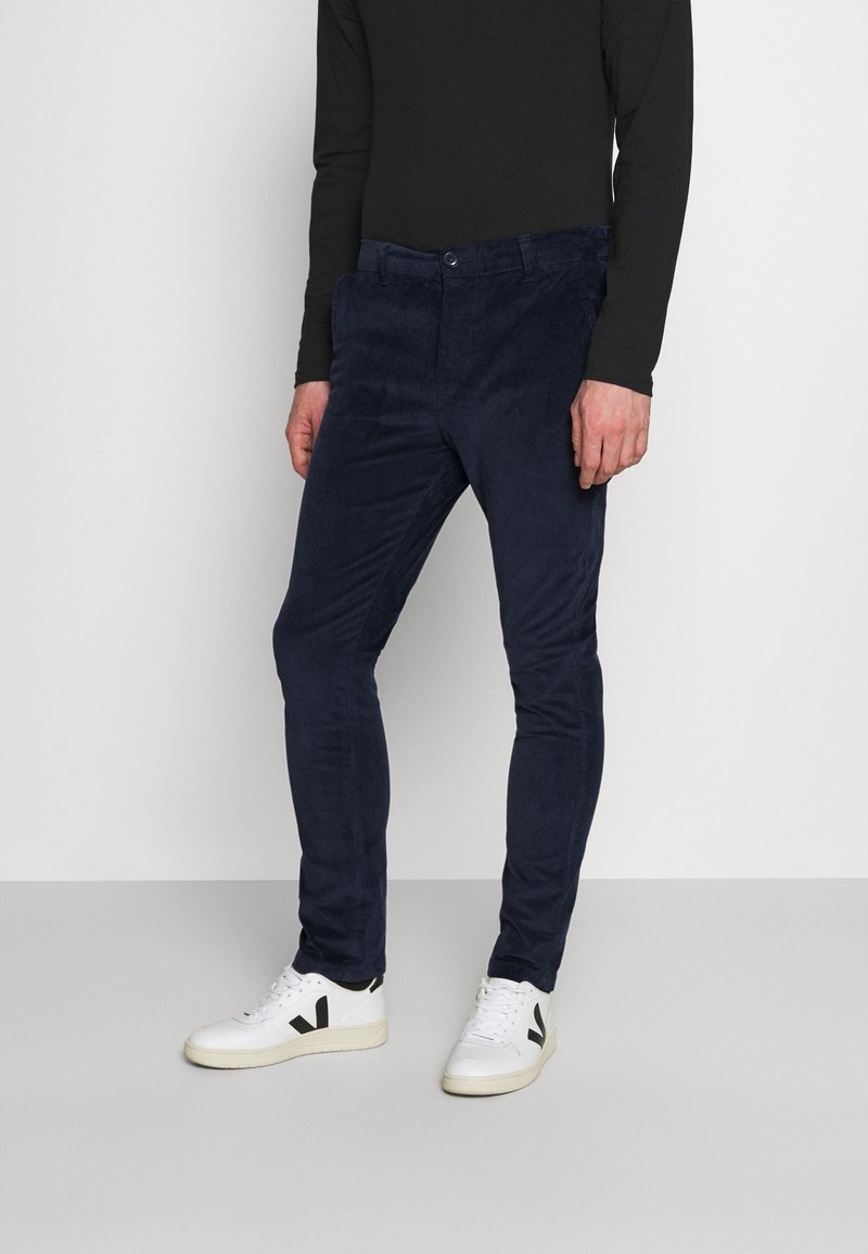 Mann trägt dunkelblaue Slim-Fit-Hose, schwarzes Langarmshirt und weiße Turnschuhe mit schwarzen Logos, steht auf grauem Boden.
