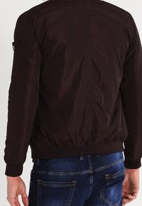 Giacca bomber marrone scuro realizzata in un tessuto leggero e morbido. Caratterizzata da polsini e vita a costine, e da una tasca con zip sulla manica sinistra.