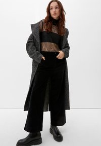 Schwarze, weit geschnittene Cordhosen kombiniert mit einem Spitzenoberteil mit langen Ärmeln, darunter ein langer grauer Fischgrätmantel. Schwarze klobige Stiefeletten.