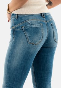 Jeans de mezclilla azul con un diseño ajustado, que cuentan con bolsillos traseros, un desvanecimiento sutil y acentos de hardware en tono dorado. Textura suave.
