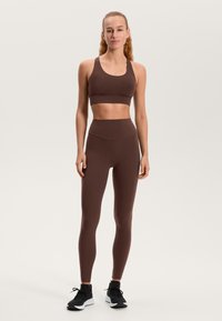 Rjavi športni modrček in legice z visokim pasom iz raztegljivega materiala. Tesno prilegajoč dizajn z minimalnimi šivi in brez vzorcev. Črne športne copate.