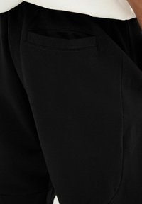 Pantalones de tela negra con una textura suave, que cuentan con un bolsillo trasero curvado que se integra en el diseño, junto con una banda elástica en la cintura.