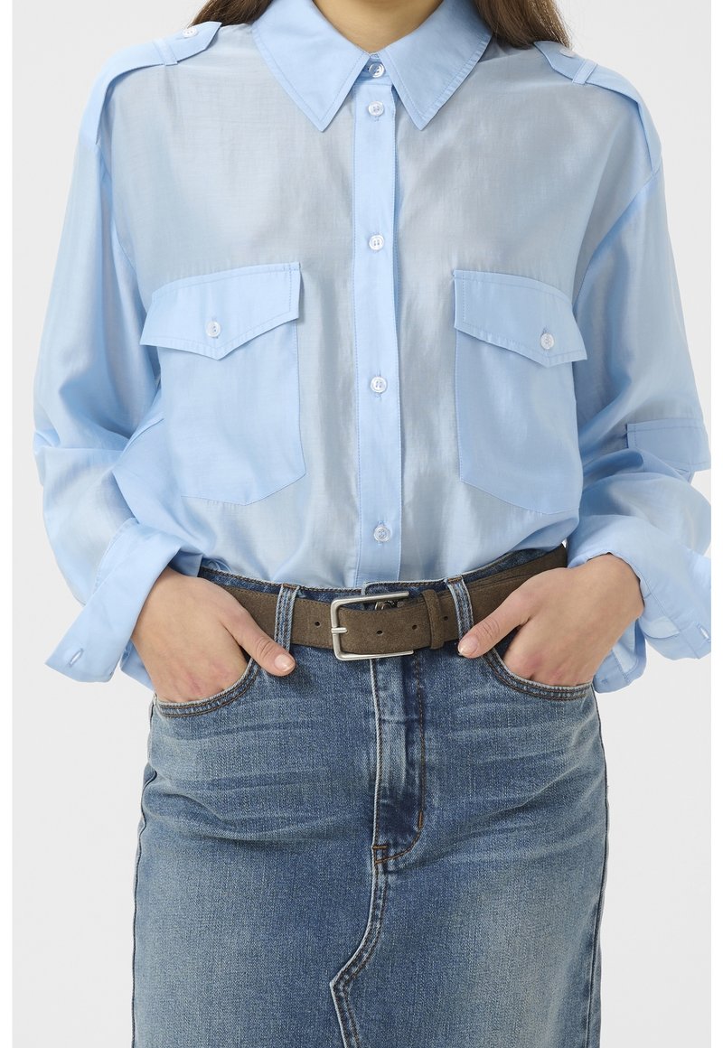Femme portant une chemise bleu clair boutonnée avec des poches poitrine, rentrée dans une jupe en jean taille haute, maintenue par une ceinture marron.