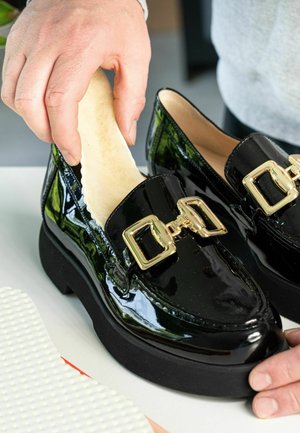 Ranka deda šviesų vidpadį į blizgantį juodą sliponą su stora padais ir auksine kvadratine saga, esančią ant balto paviršiaus.