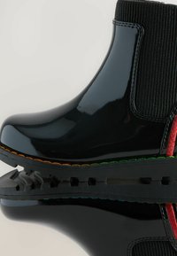 Svarta blanka ankellångstövlar med elastiska sidopaneler, kontrasterande röda och gröna detaljer, räfflad sula och dekorativ sömnad längs kanten.