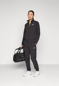 Sort træningsdragt i letvægtsstof med lynlåsjakke og elastisk talje. Hvite sneakers og sort sportstaske med logo.