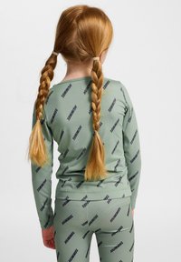 Grünes zwei-teiliges Outfit aus weichem Material, bestehend aus einem langärmligen Oberteil und Leggings mit einem wiederholten Logo-Muster in Dunkelblau.