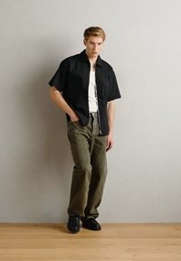 Black short-sleeve shirt over a white tank top, paired with olive green wide-leg pants and black shoes. Simple design with neutral colors.
