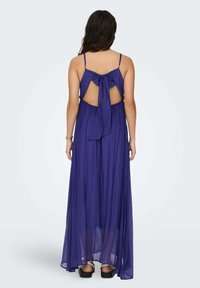 Vestido maxi en púrpura intenso, con tejido transparente y diseño de lazo en la parte posterior. Cuenta con tirantes finos y una silueta fluida que llega hasta el suelo.