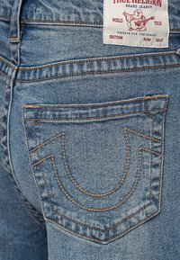 Poche arrière d'un jean en denim bleu avec surpiqûres orange et une étiquette blanche de la marque True Religion représentant une silhouette jouant de la guitare.