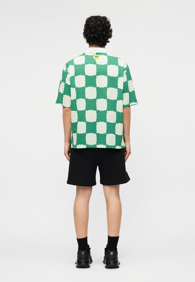 Άνδρας με σγουρά μαλλιά στέκεται με την πλάτη προς τα εμπρός, φορώντας πράσινο και λευκό καρό πουκάμισο με κοντά μανίκια, μαύρη βερμούδα, μαύρες κάλτσες και μαύρα αθλητικά παπούτσια.