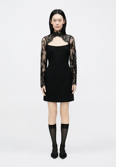 Black dress with a square neckline, long sheer lace sleeves featuring floral patterns, paired with glossy black pointed heels and knee-high stockings.