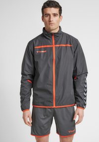 Grå sportsjakke med orange detaljer, lynlås foran og reflekterende logo. Sammen med grå shorts med kontrasterende orange kant.