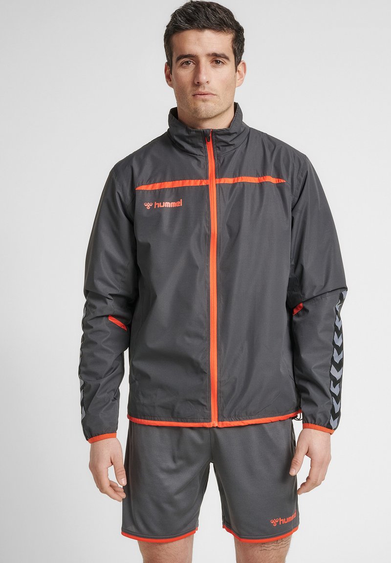 Grå sportsjakke med orange detaljer, lynlås foran og reflekterende logo. Sammen med grå shorts med kontrasterende orange kant.