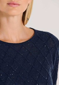 Nahaufnahme einer Person, die einen dunkelblauen strukturierten Pullover mit kleinen Pailletten und einen langen goldenen Ohrring mit schwarzen und goldenen Details trägt.
