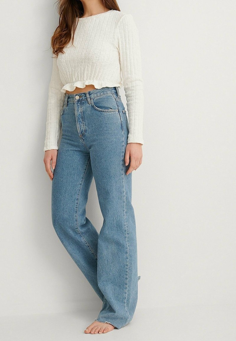 Bílý ribbovaný crop top s volánem, zkombinovaný s vysokými modrými džíny, které mají volný střih a mírně roztrhané lemy.