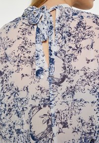 Bluza floral ușoară în nuanțe de bleumarin și alb, cu un design cu spatele închis, gât legat și o decupare în formă de cheie, textură netedă.
