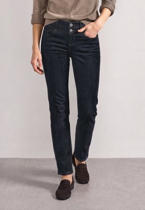 Persona que lleva pantalones azul oscuro ajustados con cierre de doble botón y mocasines marrones, de pie con una mano en el bolsillo.