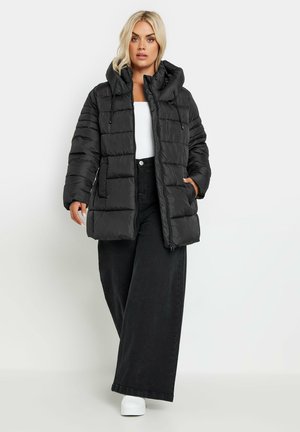 Sort puffervest med hætte, med lynlåslukning, vandret quiltet design og to sidelommer. Bæres over en hvid top og sorte jeans.