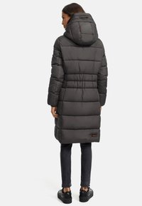 Geacă puf în gri închis, cu un design matlasat, de lungime medie, cu glugă ajustabilă și buzunare laterale, asociată cu pantaloni negri și adidași.