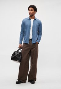 Camisa azul abotoada sobre uma t-shirt branca, combinada com calças largas castanhas e uma mala preta texturizada. Sapatos pretos completam o conjunto.