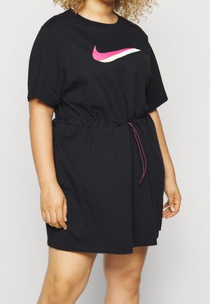 Osoba ubrana w czarną sukienkę z krótkim rękawem, opinającą talię i różowe logo Nike na piersi.