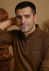 Pull en demi-zip marron en tissu texturé, avec des accents côtelés et un petit logo bleu de la marque sur la poitrine.