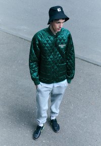 Chaqueta bomber acolchada verde con bolsillos, pantalones de chándal grises y sombrero de cubo negro. Zapatillas negras con acentos amarillos. Entorno urbano al aire libre.