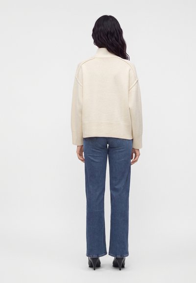 Femme aux longs cheveux foncés portant un pull à col roulé crème, un jean droit bleu et des bottines noires à talons, debout de dos.