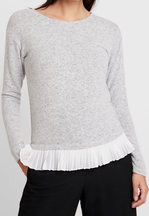 Kvinde iført en lysegrå langærmet sweater med en hvid plisseret kant, kombineret med sorte bukser, stående med hænderne i lommerne.