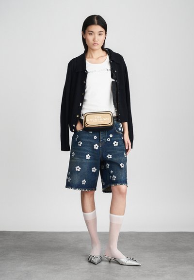 Kvinne med en svart cardigan over hvit T-skjorte, dongerishorts med hvite blomster mønster, hvite gjennomsiktige sokker, sølvfargede kitten heels, og en lys brun crossover bag.
