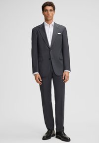 Mężczyzna stoi ubrany w ciemnoszary, dopasowany garnitur, biały koszulę, czarne buty wizytowe i biały poszetkę.