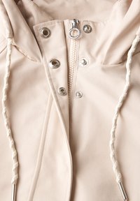 Veste beige clair avec fermeture éclair frontale, quincaillerie en métal et capuche réglable. Tissu lisse, résistant à l'eau, avec des coutures renforcées.