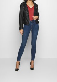 Giubbotto biker in pelle nera, top rosso a scollo a V e jeans attillati blu scuro abbinati a tacchi neri a punta; texture morbide e design aderente.