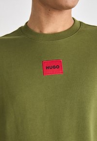 Homme portant un t-shirt vert olive à col rond avec un petit écusson rouge libellé "HUGO" centré sur la poitrine.