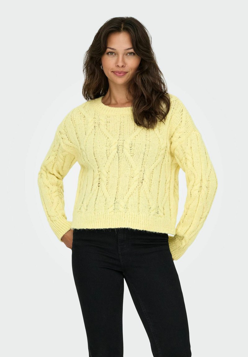 Pull en mailles jaunes avec un motif texturé et des manches côtelées. Associé à un jean noir, le pull présente un col rond et une coupe décontractée.