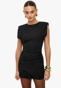 Robe mini noire à manches courtes structurées, détails froncés sur le côté et texture lisse, accessoirisée avec des bracelets dorés.