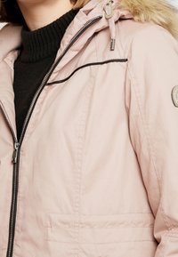 Jasnoróżowa ocieplana kurtka z kapturem wyłożonym futerkiem, przednim zamkiem błyskawicznym i czarnymi detalami z lamówki. Posiada boczne kieszenie oraz teksturowany kołnierz.
