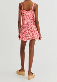 Frau in einem pinkfarbenen, blumenbedruckten, ärmellosen Mini-Kleid und hellbraunen, geflochtenen Slipper-Plattformsandalen, die vor einem schlichten weißen Hintergrund steht.