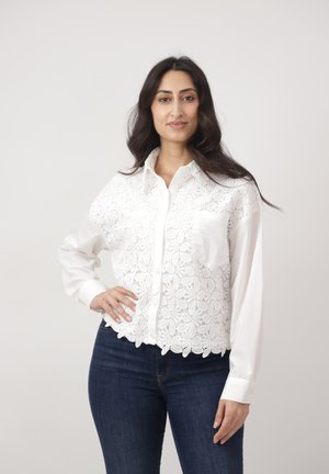 Kobieta o długich ciemnych włosach, ubrana w białą koronkową bluzkę zapinaną na guziki i ciemnoniebieskie dżinsy, stoi z jedną ręką na biodrze.