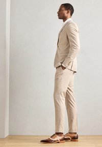Completo su misura beige chiaro con una vestibilità slim, abbinato a scarpe eleganti bicolore marroni e crema con perforazioni decorative.