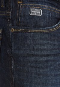Mørkeblå denimjeans med en jevn tekstur, gule sømmer, en metalltag på linningen, og en frontlomme med en nittedetalj.