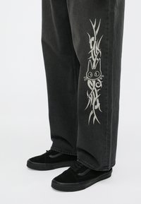 Pantaloni di denim nero con motivo grafico bianco raffigurante un gatto e motivi astratti a spirale lungo il lato, abbinati a scarpe da ginnastica nere.